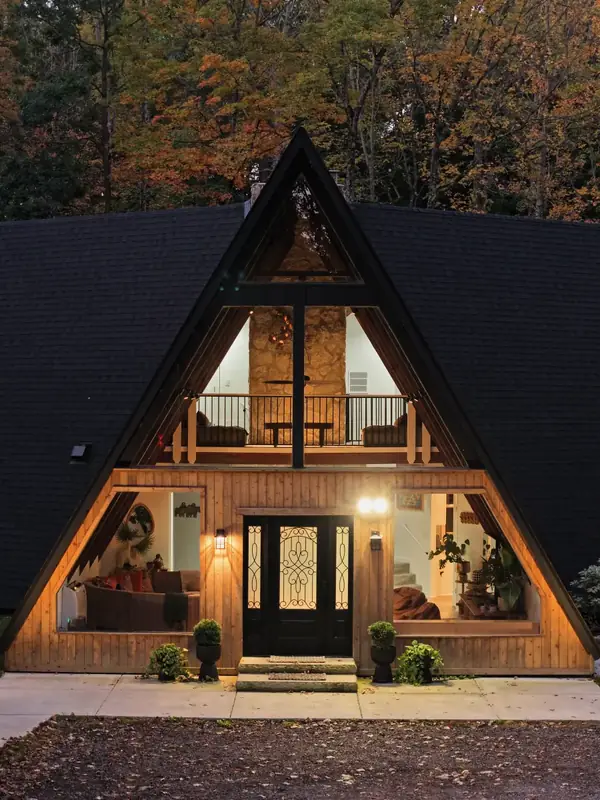 Glass front of an A-frame cabin at dusk showing a lit loft, stone wall and living room at Parker Reserve, Ohio