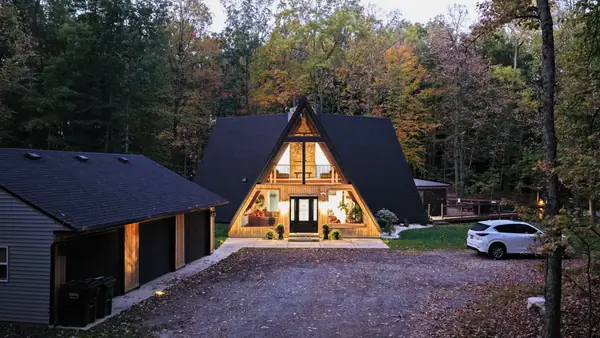 A-frame cabin lit from inside at dusk, set in fall woods beside a garage and gravel drive at Parker Reserve, Ohio